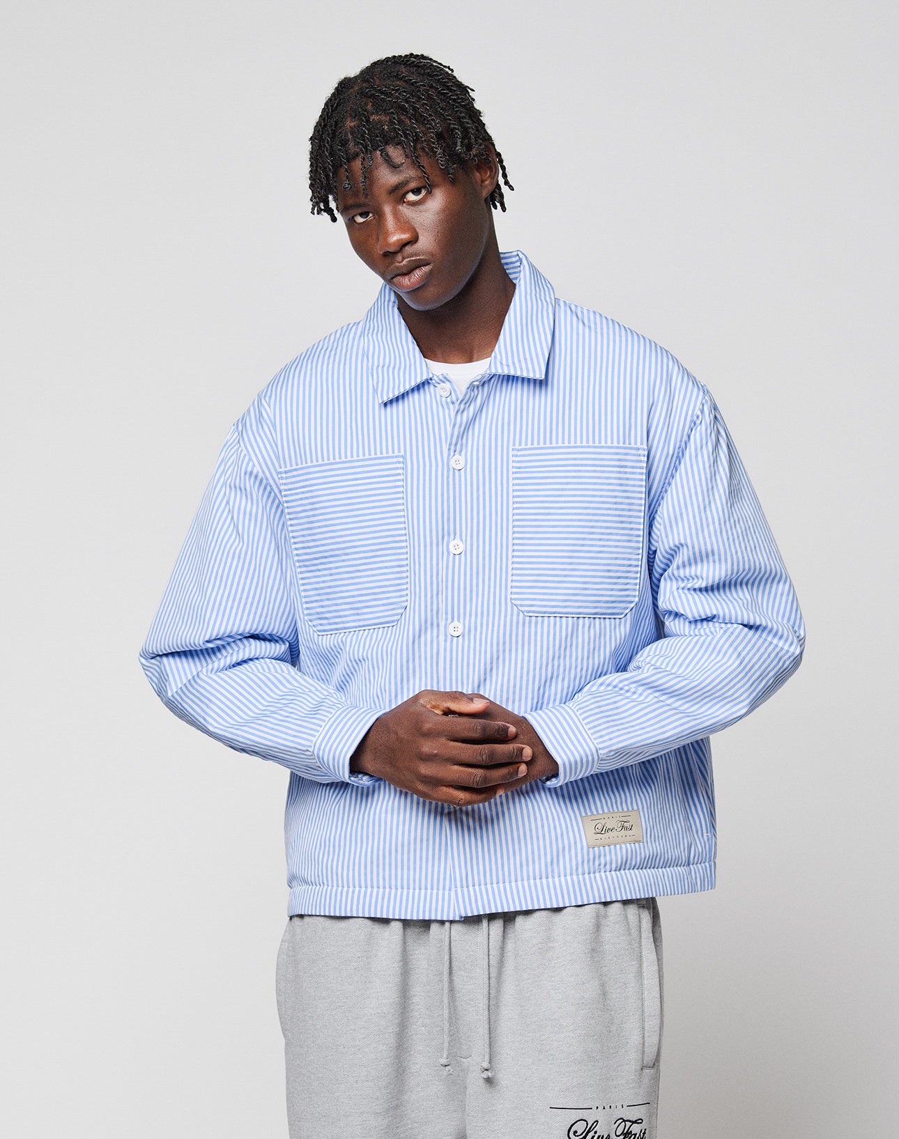 A young man with short, twisted hair stands against a plain background wearing the LIVE FAST DIE YOUNG LFDY Striped Padded Shirt and light gray sweatpants. His hands are clasped in front of him.