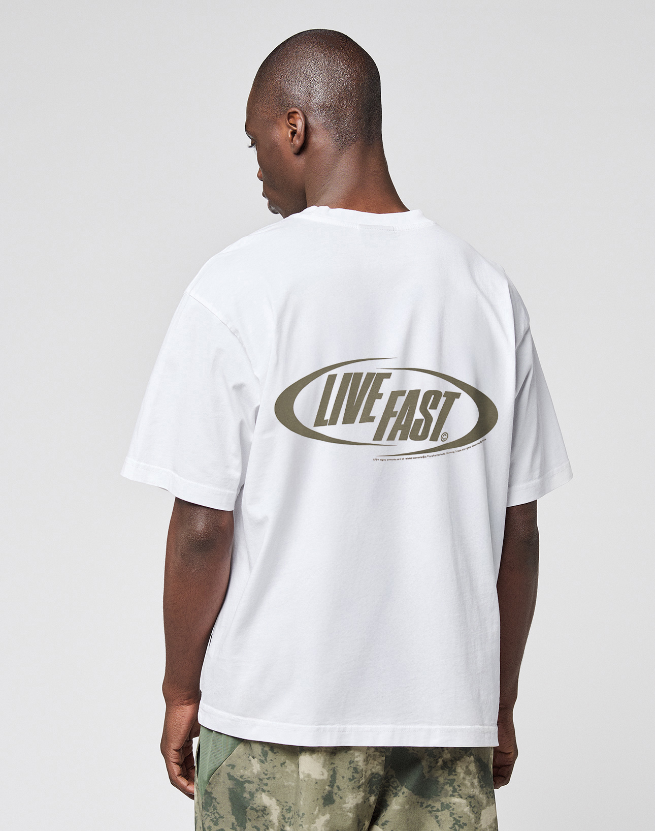 A person models the LFDY Sphere Tee by LIVE FAST DIE YOUNG—an oversized white cotton shirt featuring “LIVE FAST.” in a bold oval on the back. Pairs well with green and beige camo pants.
