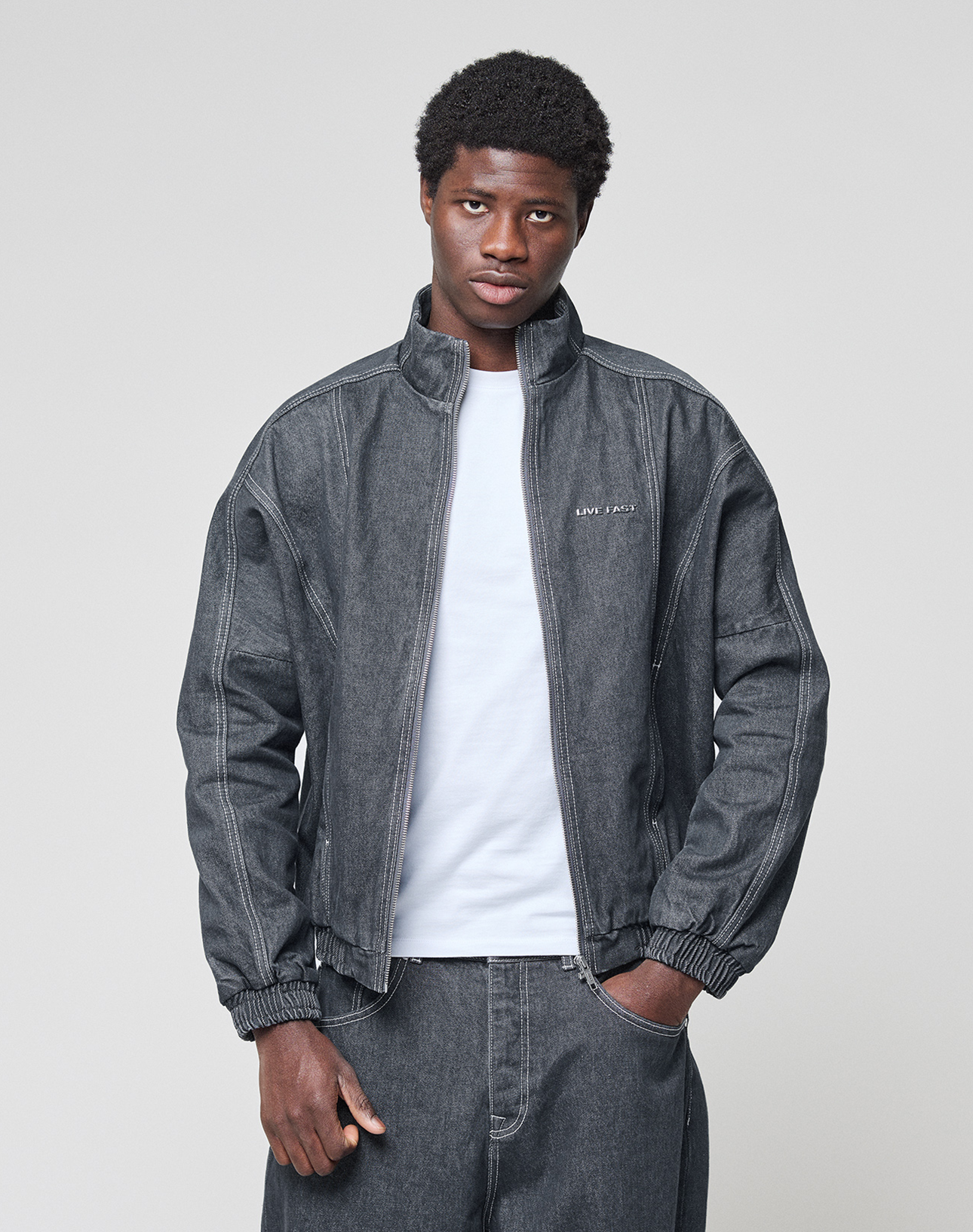 A person wears the LFDY Denim Trackjacket by LIVE FAST DIE YOUNG in dark gray over a white t-shirt and matching denim pants, standing against a plain light gray background and looking at the camera with a neutral expression.