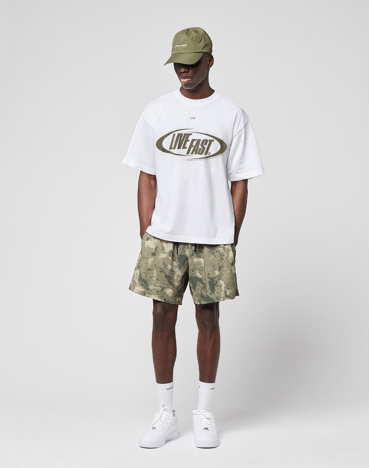 A person stands against a plain background wearing a green cap, LIVE FAST DIE YOUNG LFDY Sphere Tee (oversize fit, white with front and back print), camouflage shorts, white socks, and white sneakers, hands in pockets facing forward.
