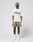 A person stands against a plain background wearing a green cap, LIVE FAST DIE YOUNG LFDY Sphere Tee (oversize fit, white with front and back print), camouflage shorts, white socks, and white sneakers, hands in pockets facing forward.
