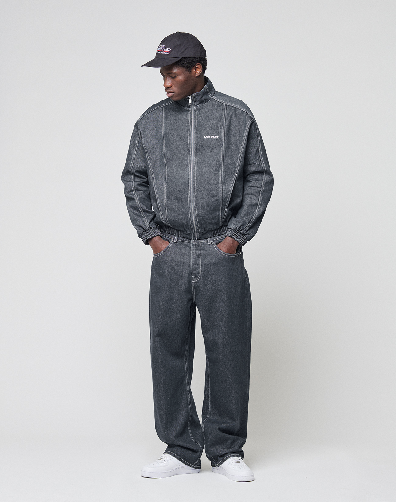 Wearing the LIVE FAST DIE YOUNG LFDY Baggy Denim set in dark gray 100% Baumwolle, a black baseball cap, and white sneakers, a person stands against a plain light background with hands in pockets, looking to the side.