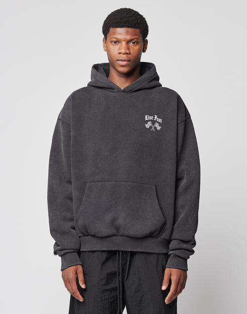A young man stands facing forward, wearing the Ride or Die Hooded by LIVE FAST DIE YOUNG—a black oversize fit hoodie with a small white Live Fast graphic on the left chest, doppellagige Kapuze, and black pants—against a light gray background.