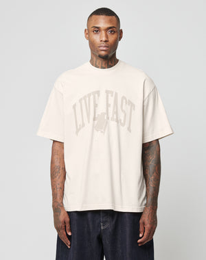 A tattooed man wears the LIVE FAST DIE YOUNG Live Fast Ice Tee, a loose beige Baumwolle t-shirt in Oversize Fit with a LIVE FAST print and popsicle graphic. He stands facing forward against a plain gray background, arms relaxed.