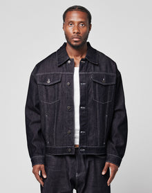 A man with braided hair wears an oversize LFDY Denim Jacket by LIVE FAST DIE YOUNG and matching raw-denim jeans over a white shirt, standing against a plain light gray background.