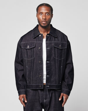 A man with braided hair wears an oversize LFDY Denim Jacket by LIVE FAST DIE YOUNG and matching raw-denim jeans over a white shirt, standing against a plain light gray background.