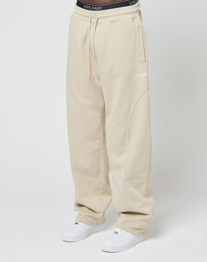 A person wears the LIVE FAST DIE YOUNG Basic Wide Jogger in beige with an elastic waistband and LIVE FAST embroidery, paired with white sneakers, standing against a plain white background.