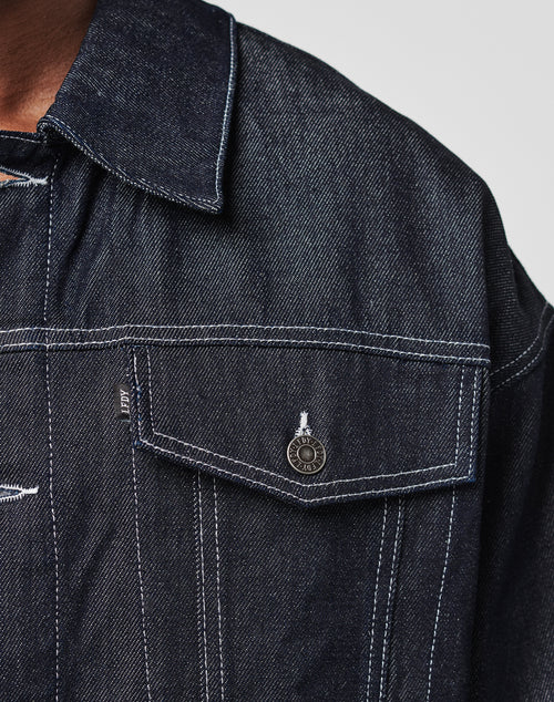 Close-up of the LFDY Denim Jacket by LIVE FAST DIE YOUNG in dark raw denim, with white stitching, a pointed collar, and a flap chest pocket featuring an RBY-engraved metallic button. The texture and weave are visibly detailed.