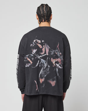 A person with braided hair faces away, wearing the LIVE FAST DIE YOUNG LFDY Canine Longsleeve—an oversize-fit black shirt made from 100% cotton, featuring three barking Dobermans on the back and light stripes on the sleeves.