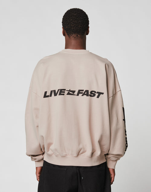 A person with short curly hair, seen from behind, wears the LIVE FAST DIE YOUNG Live For It Sweater—an oversized beige sweatshirt with LIVE FAST in bold black on the back. Made from 100% Baumwolle for comfort against a plain background.