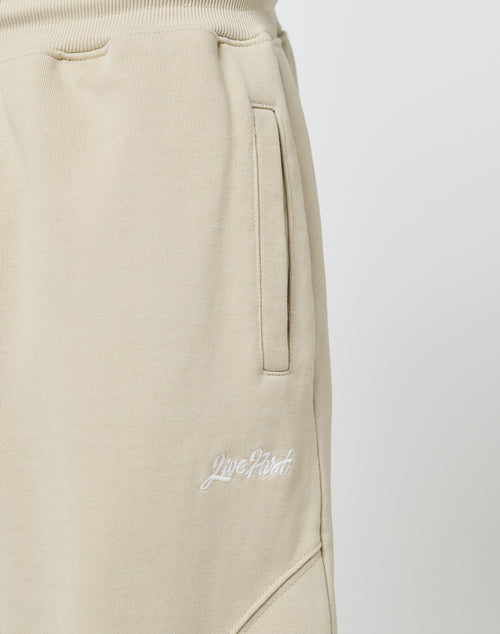 Close-up of Basic Wide Jogger sweatpants by LIVE FAST DIE YOUNG in beige, featuring a wide fit, side pockets, elastic waistband, and white Live Fast embroidery near the hip. Made from soft cotton. Plain light background.