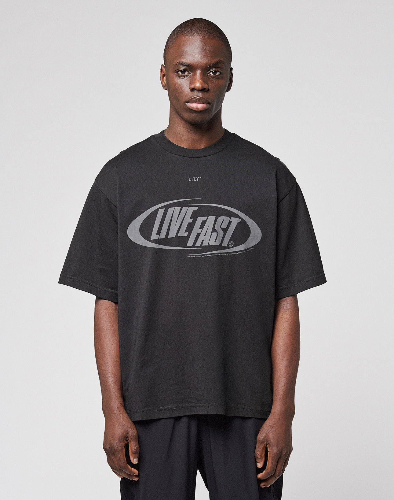 A person wearing the LFDY Sphere Tee by LIVE FAST DIE YOUNG—an oversized black cotton T-shirt with "LIVE FAST." boldly printed on the front—stands against a plain, light gray background.
