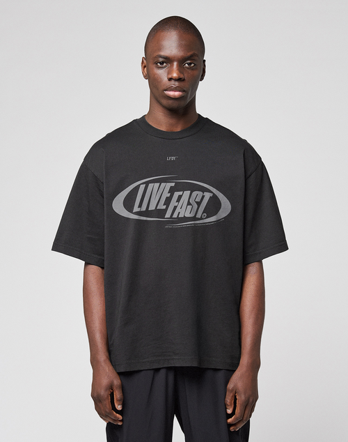 A person wearing the LFDY Sphere Tee by LIVE FAST DIE YOUNG—an oversized black cotton T-shirt with "LIVE FAST." boldly printed on the front—stands against a plain, light gray background.