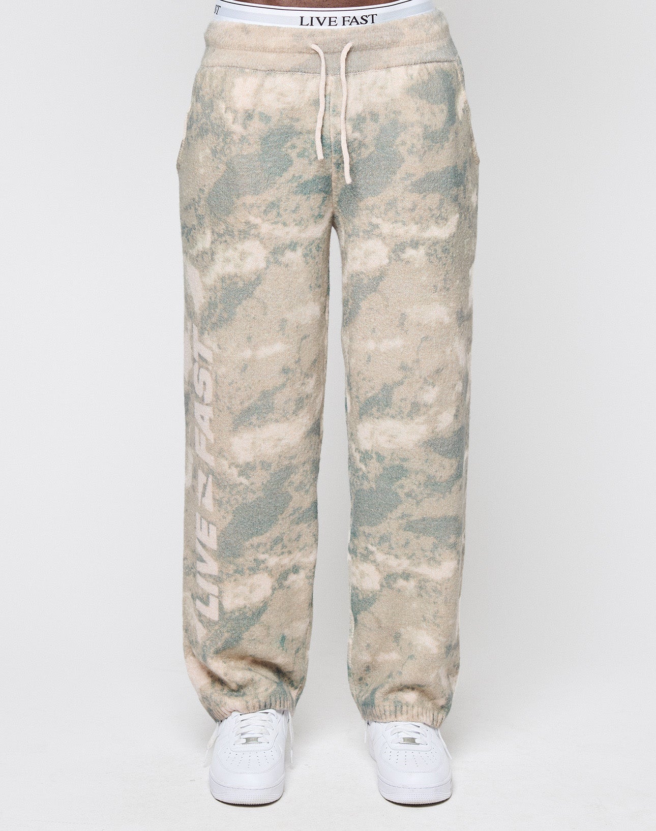 A person wearing LIVE FAST DIE YOUNG’s Live Fast Boucle Pants in light green and beige tie-dye with a broad elastic waistband, white drawstrings, and ‘LIVE FAST’ vertically on one leg, paired with white sneakers against a plain background.