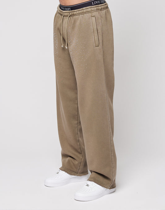 A person wears the LIVE FAST DIE YOUNG Live Fast Rhinestone Jogger in tan with rhinestone logo detail and a visible black waistband that reads "LIVE FAST," paired with white sneakers, standing against a plain white background.