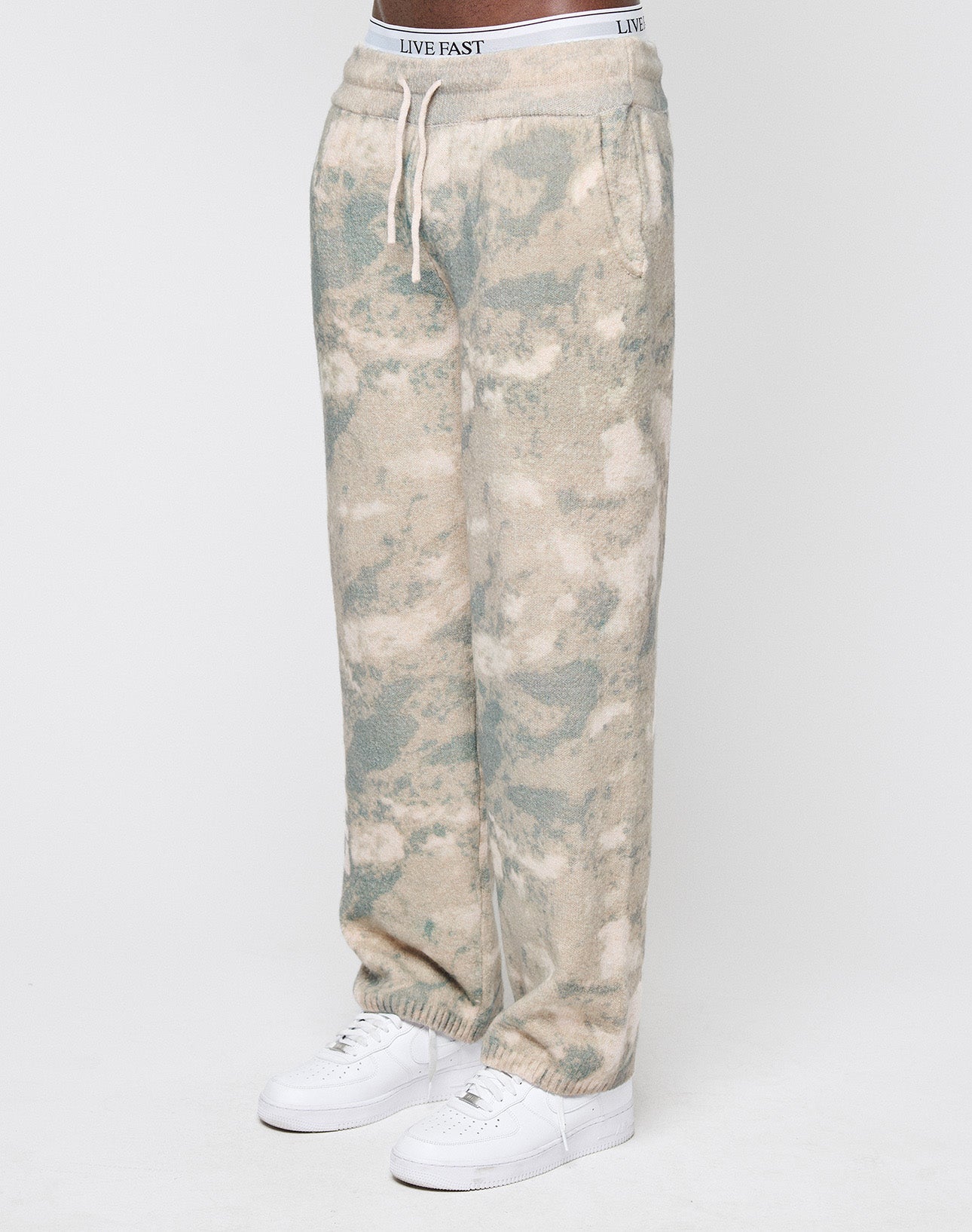 A person wears LIVE FAST DIE YOUNG's Live Fast Boucle Pants in relaxed beige and light blue tie-dye with a wide elastic waistband, white drawstring, and visible LIVE FAST branding—styled with white sneakers for a comfy, oversized look.