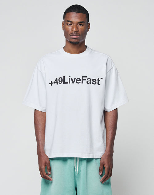 A man wears the LIVE FAST DIE YOUNG Basic +49 Tee, an oversize fit white T-shirt with a bold black +49LiveFast front print, made from 100% Baumwolle. He pairs it with light green shorts and stands against a light gray background.