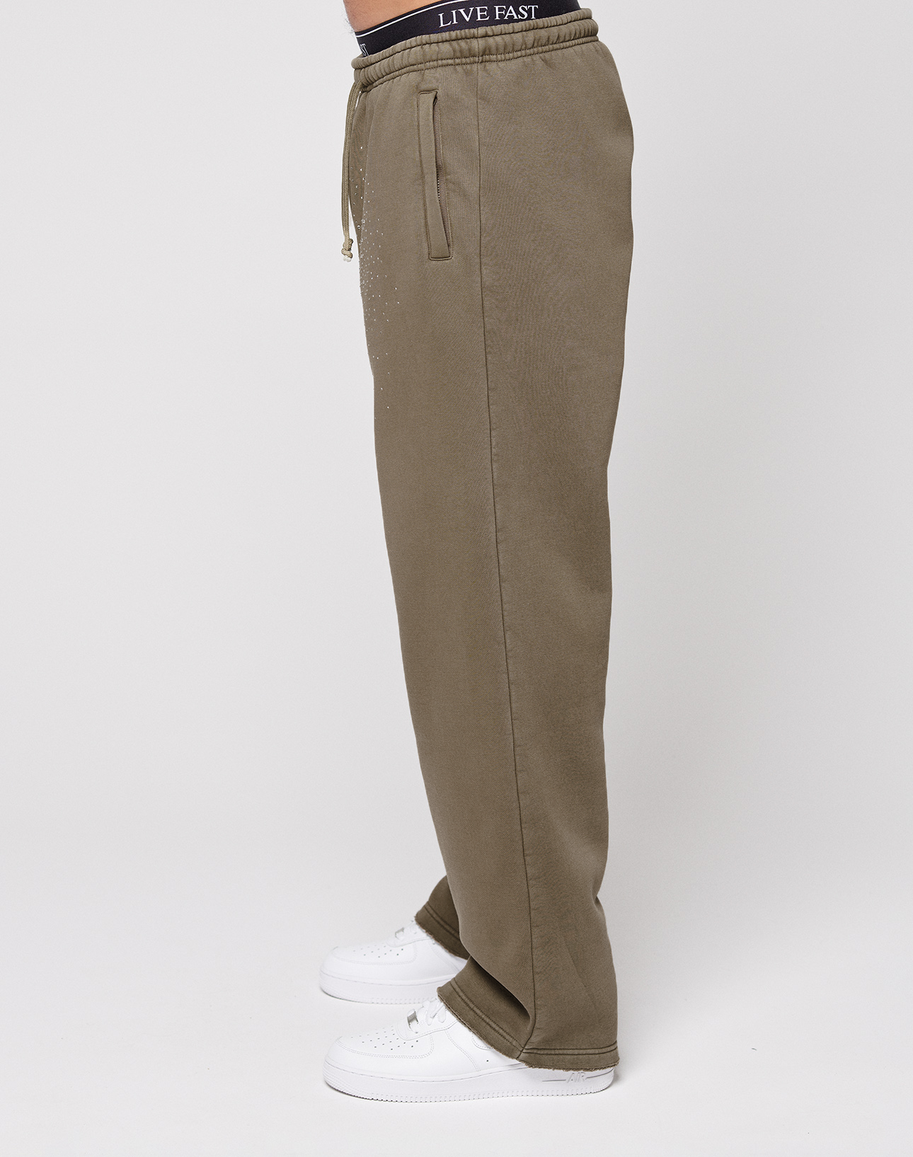 A person in LIVE FAST DIE YOUNG’s olive green Live Fast Rhinestone Jogger with side pockets and an oversize fit, white sneakers, and black "LIVE FAST" waistband underwear, shown in side profile against a plain white background.