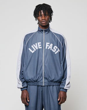 A man with medium-length curly hair wears a blue and gray LFDY Arch Trackjacket by LIVE FAST DIE YOUNG, featuring LIVE FAST printed in white on the front. He stands facing forward with hands in the side pockets against a plain light background.