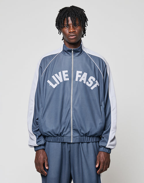 A man with medium-length curly hair wears a blue and gray LFDY Arch Trackjacket by LIVE FAST DIE YOUNG, featuring LIVE FAST printed in white on the front. He stands facing forward with hands in the side pockets against a plain light background.