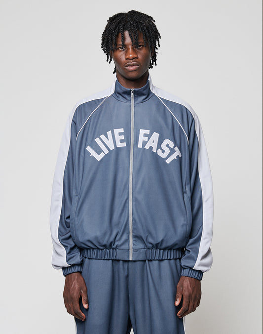 A man with short dreadlocks wears the LFDY Arch Trackjacket by LIVE FAST DIE YOUNG, featuring blue and gray colors, LIVE FAST across the chest, and elastische Bündchen, against a plain light background.