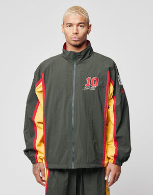 A person with short blond hair wears a LIVE FAST DIE YOUNG Westend Trackjacket in dark green, red, and yellow polyester with multi-logo embroidery, standing against a plain light gray background.