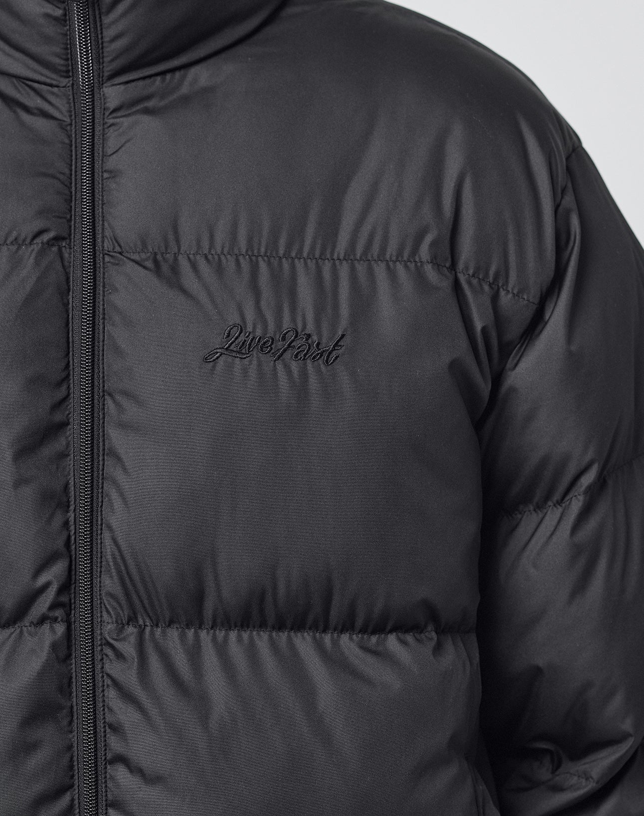 Close-up of a person wearing the black LFDY Puffer Jacket by LIVE FAST DIE YOUNG with an oversize fit and "Live Fast" logo stitching on the chest. Only the upper torso is visible.