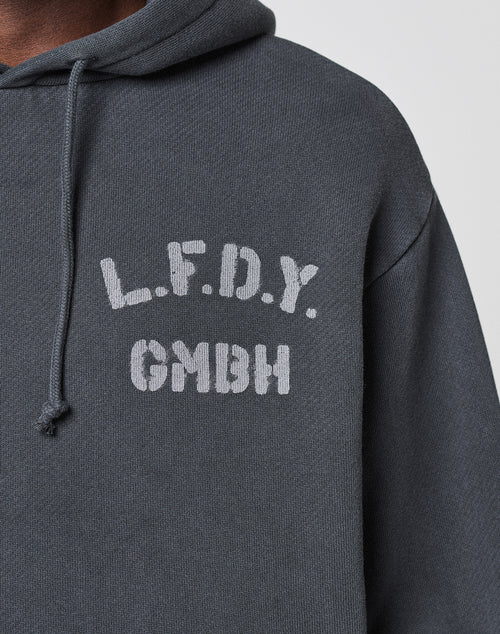 A close-up of the LFDY GmbH Hooded by LIVE FAST DIE YOUNG, a dark gray oversized hoodie with white "L.F.D.Y. above GMBH" text on the left chest and a visible drawstring on the left side of the hood.