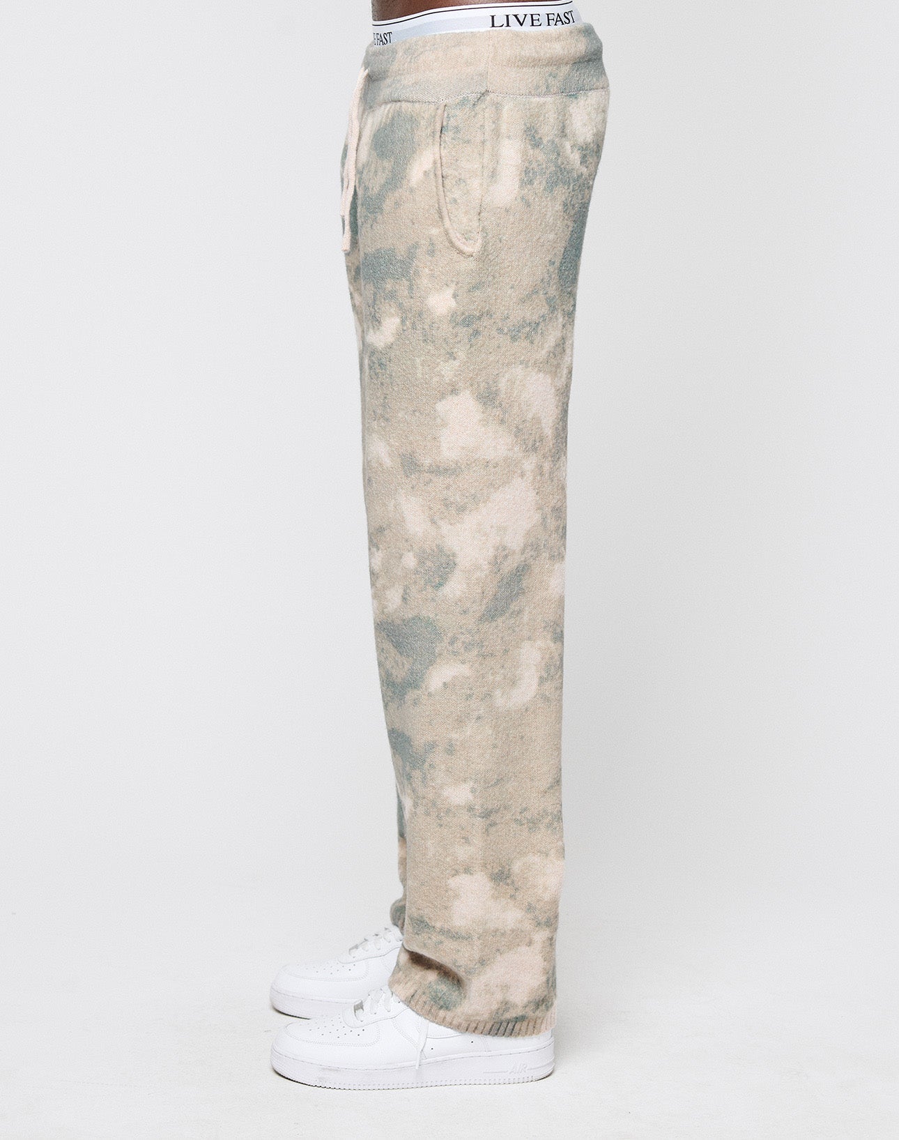 A person in Live Fast Boucle Pants by LIVE FAST DIE YOUNG, featuring tan and green tie-dye, a breiter Elastikbund, wide fit, and gerades Bein with "LIVE FAST" on the waistband, stands in profile with white sneakers against a white background.