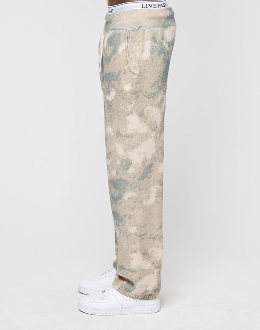 A person in Live Fast Boucle Pants by LIVE FAST DIE YOUNG, featuring tan and green tie-dye, a breiter Elastikbund, wide fit, and gerades Bein with "LIVE FAST" on the waistband, stands in profile with white sneakers against a white background.