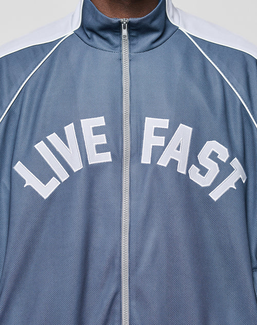 A person is wearing the LFDY Arch Trackjacket by LIVE FAST DIE YOUNG, a blue zip-up jacket with white piping, elastic cuffs, and LIVE FAST printed in bold white letters on the chest.