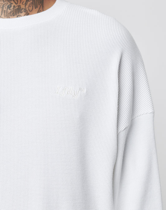 A close-up of a person wearing the LIVE FAST DIE YOUNG Basic Waffle Longsleeve, an oversize white 100% Baumwolle sweatshirt with subtle chest Logo-Stick embroidery. Part of their neck tattoo appears above the collar.