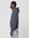 A person with short, braided hair stands in profile wearing a dark gray LIVE FAST DIE YOUNG LFDY GmbH Hooded sweatshirt and light blue jeans against a plain white background.
