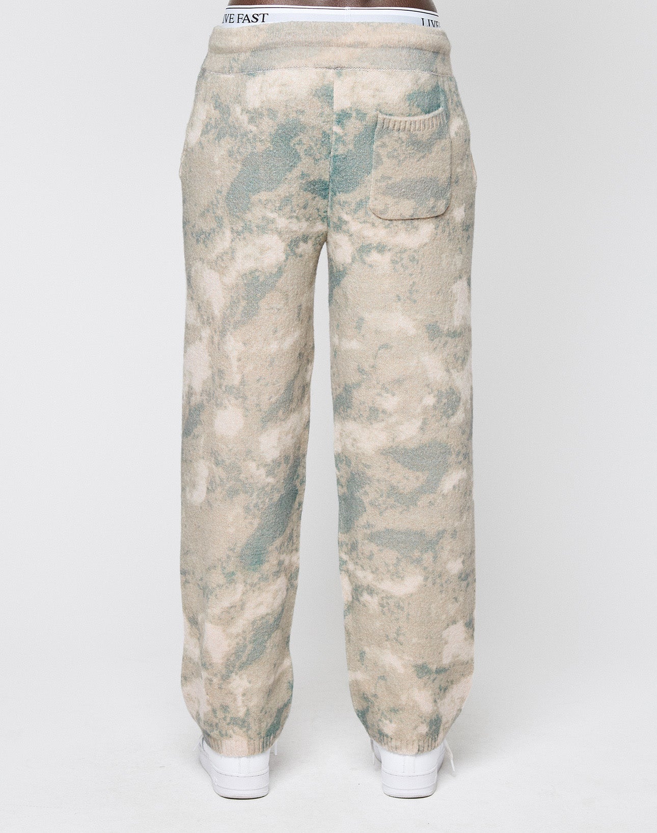A person seen from the back wears LIVE FAST DIE YOUNG’s Live Fast Boucle Pants in beige and green tie-dye with a wide elastic waistband, straight leg, single rear pocket, and white shoes, set against a plain white background.