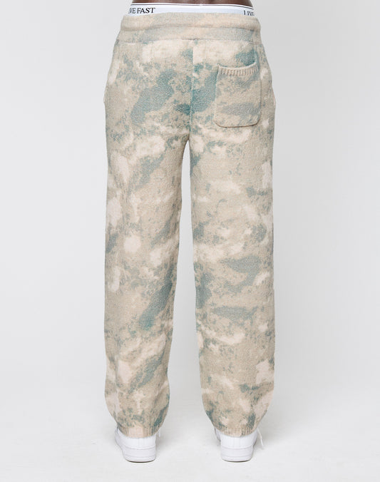 A person seen from the back wears LIVE FAST DIE YOUNG’s Live Fast Boucle Pants in beige and green tie-dye with a wide elastic waistband, straight leg, single rear pocket, and white shoes, set against a plain white background.
