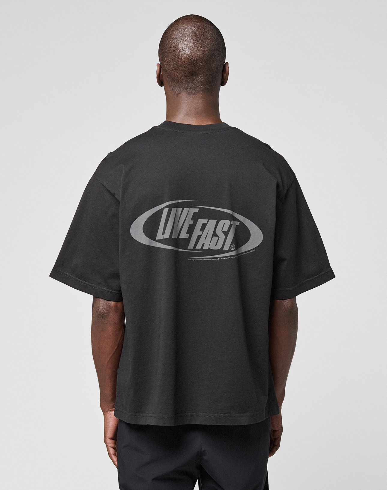 A person stands with their back to the camera against a plain light gray background, wearing the LIVE FAST DIE YOUNG LFDY Sphere Tee—an oversize black Baumwolle t-shirt featuring a bold "LIVE FAST!" backprint.