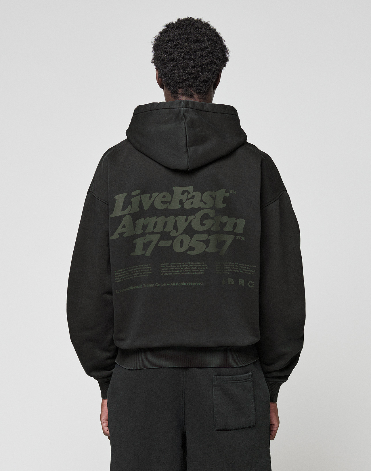 A person wearing the LIVE FAST DIE YOUNG Basic 360 Hooded black hoodie with the hood up, seen from behind. The back showcases large green "LiveFast ArmyGrn 17-0517" text and small writing—a stylish front and back print design.