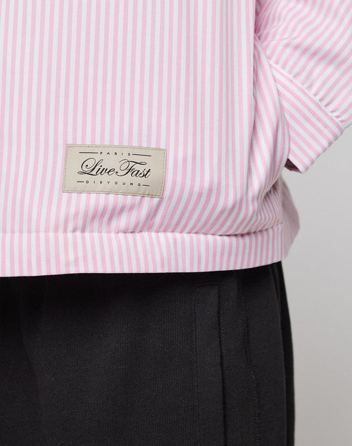 A close-up of a person in the LFDY Striped Padded Shirt by LIVE FAST DIE YOUNG, featuring pink and white vertical stripes and a "Live Fast" patch near the hem, paired with black pants; their right arm is bent at the elbow.