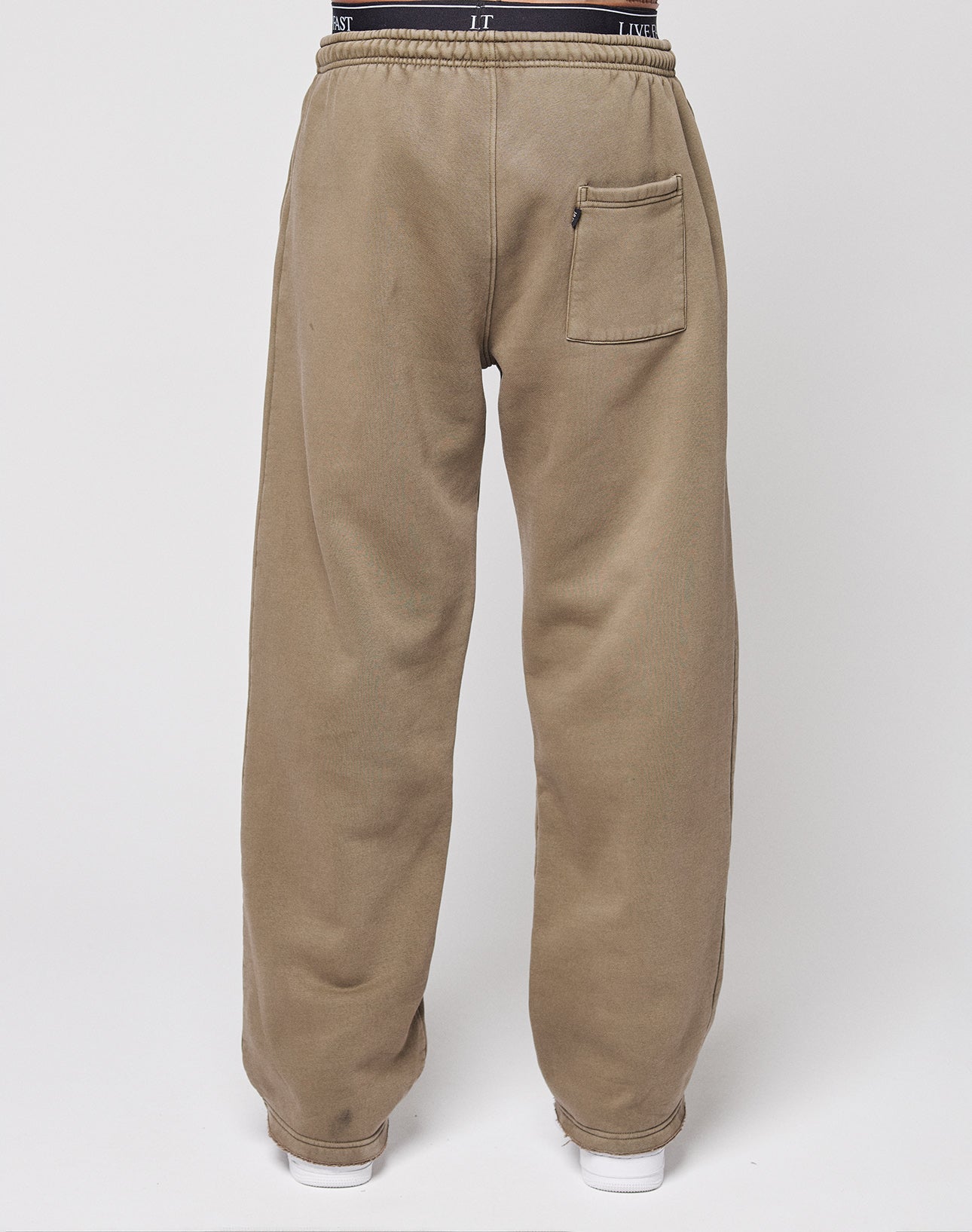 A person stands with their back to the camera, wearing LIVE FAST DIE YOUNG's Live Fast Rhinestone Jogger—light brown, loose-fit sweatpants with a vintage wash, elastic waistband, and single back pocket—against a plain white background.