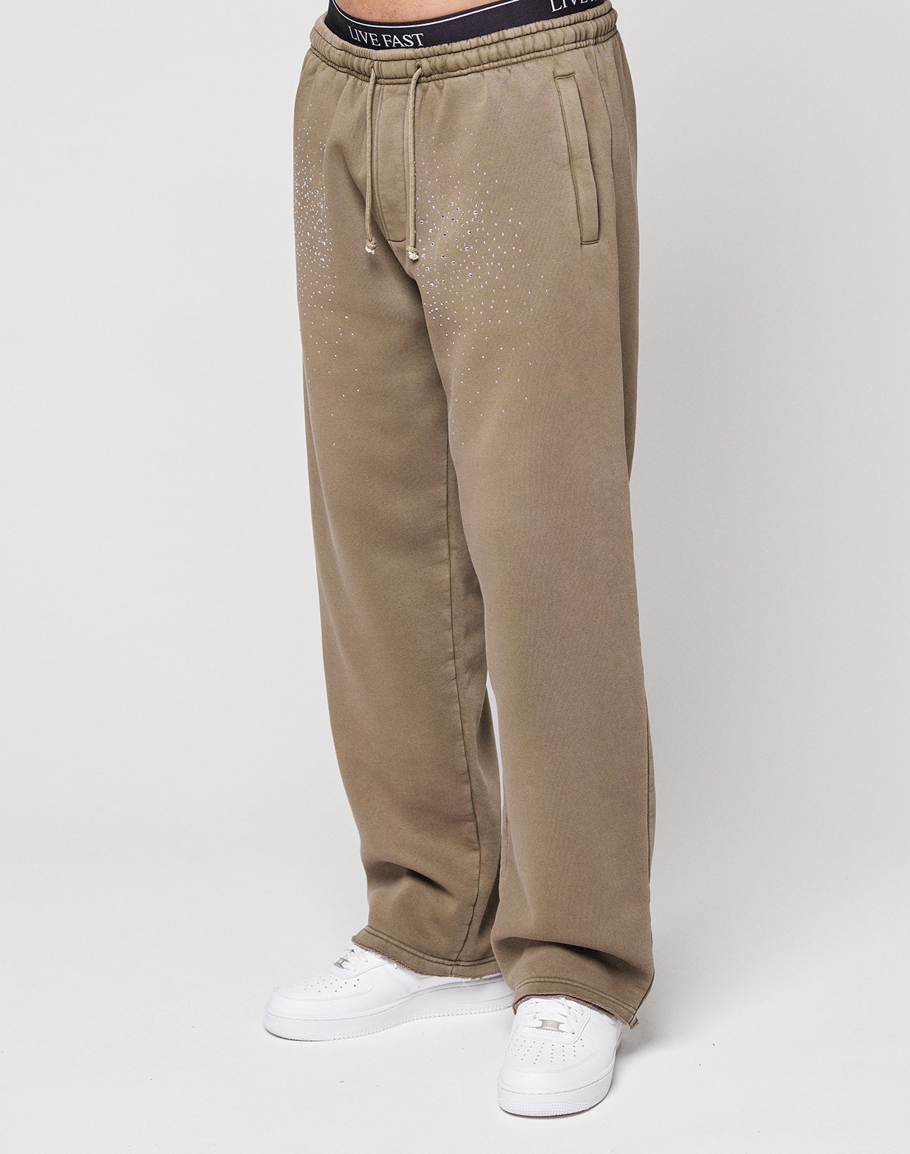 Someone stands against a plain white background wearing the LIVE FAST DIE YOUNG Live Fast Rhinestone Jogger—loose-fitting, tan sweatpants featuring a rhinestone logo, small studs, and a visible waistband with "LIVE FAST" printed on it.