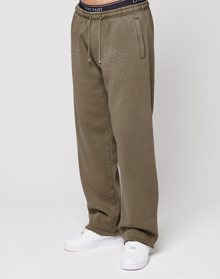 A person wears the LIVE FAST DIE YOUNG Live Fast Rhinestone Jogger—loose-fitting olive green sweatpants with a vintage wash, rhinestone dot accents on the upper thighs, and a "LIVE FAST" waistband. Paired with white sneakers against a plain white background.