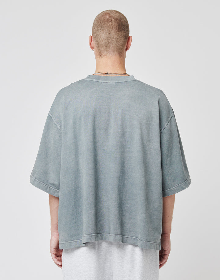 A person with short hair, facing away from the camera, wears a faded blue-gray Cropped Oversize Tee by LIVE FAST DIE YOUNG and light pants against a plain white background.