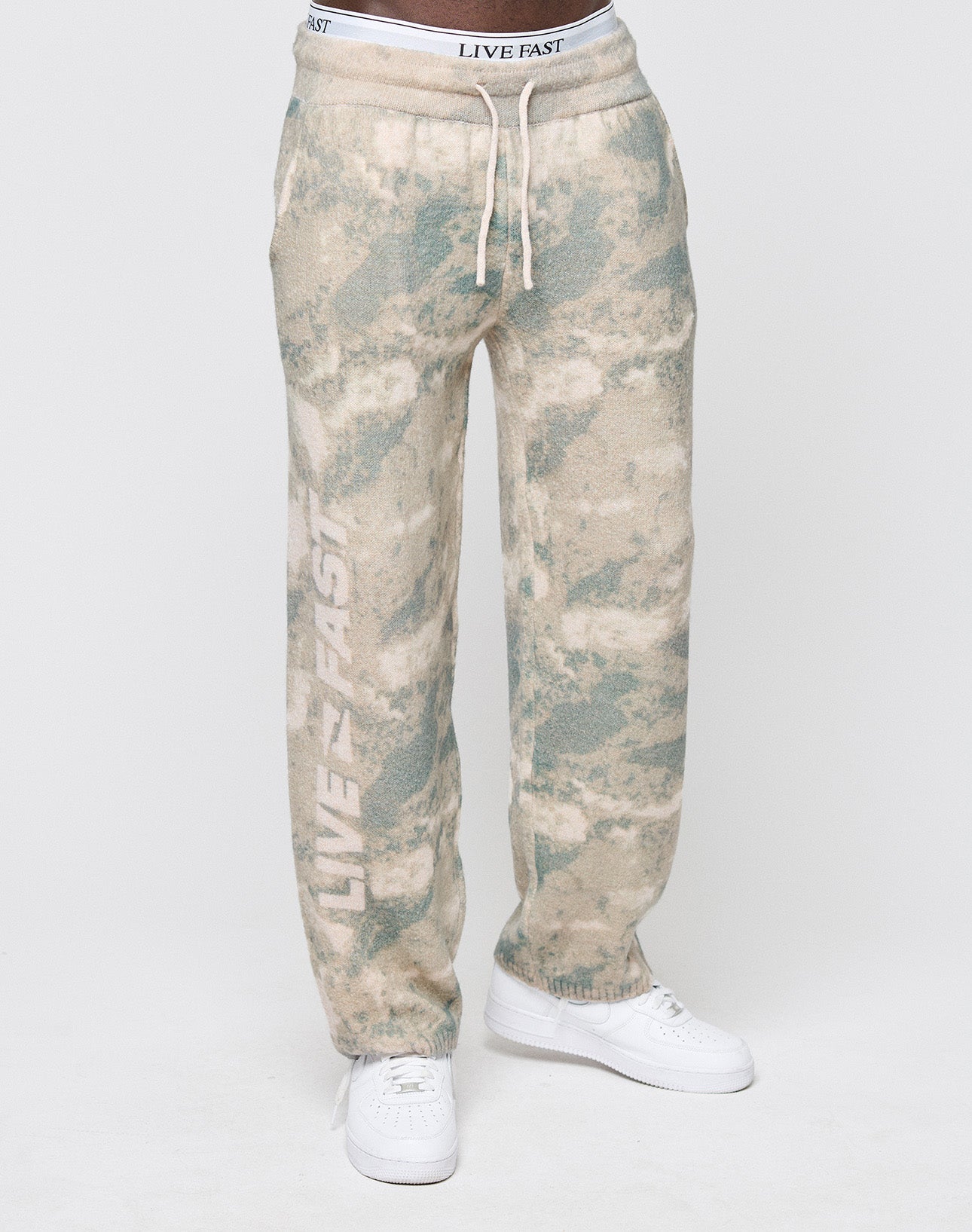 Someone wears LIVE FAST DIE YOUNG's Live Fast Boucle Pants—light-colored, tie-dye sweatpants with a wide elastic waistband and "LIVE FAST" printed vertically on one leg, styled with white sneakers against a plain white background.