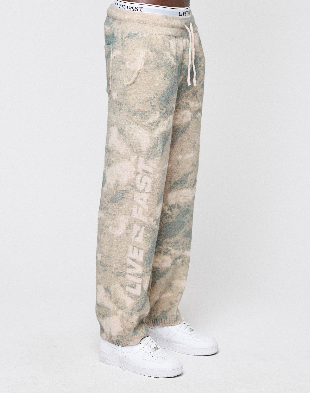 A person wears LIVE FAST DIE YOUNG’s Live Fast Boucle Pants in beige and green tie-dye, featuring a wide elastic waistband, straight leg, and LIVE FAST printed vertically on the left leg, styled with white sneakers against a plain background.
