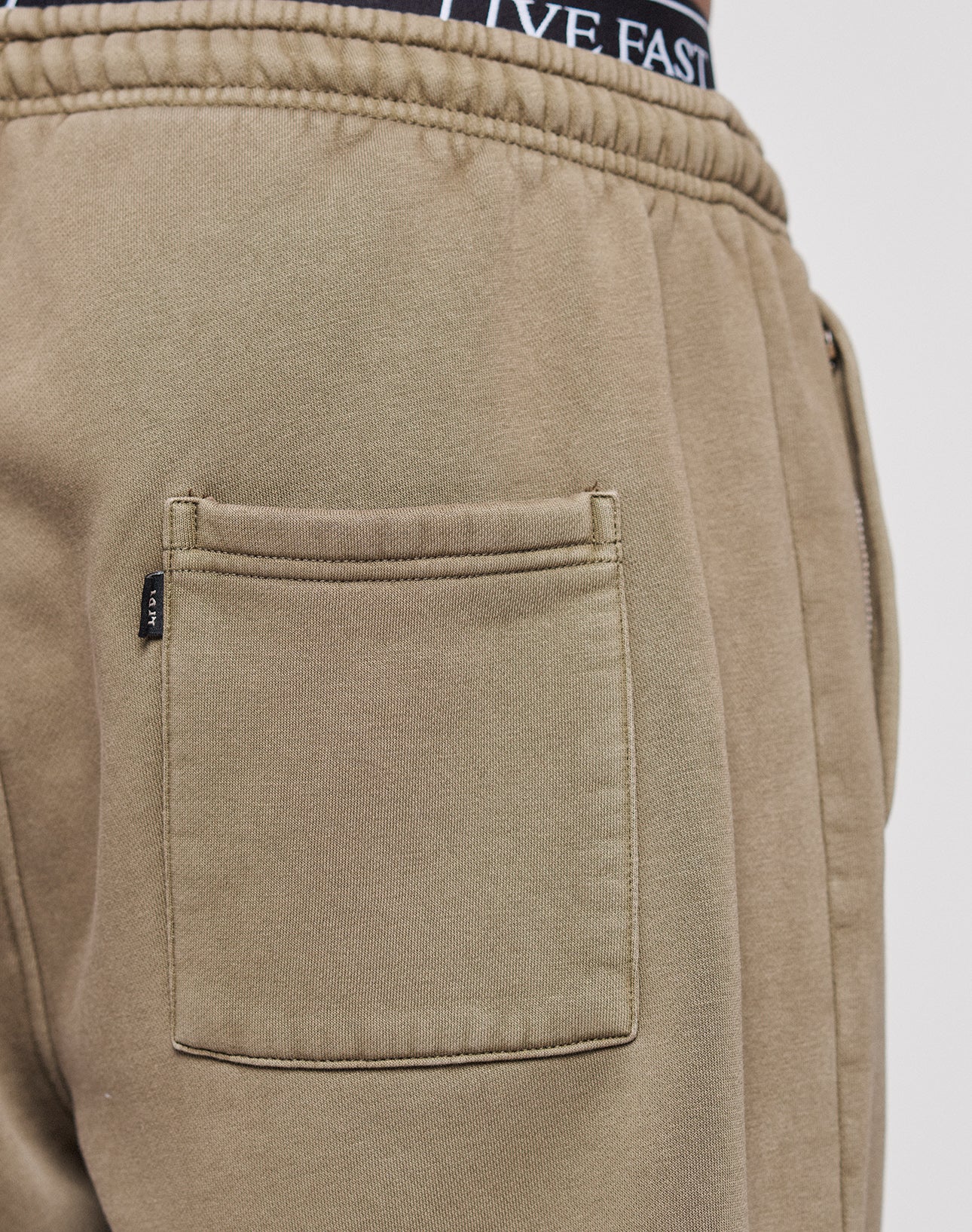 Close-up of a person wearing LIVE FAST DIE YOUNG's Live Fast Rhinestone Jogger, featuring an elastic waistband, back pocket, and subtle vintage wash; black underwear with a visible LIVE FAST waistband is partially shown.