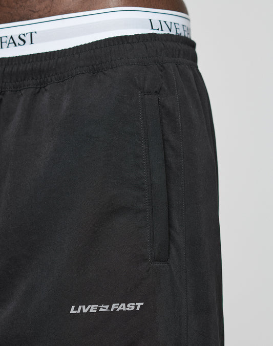 Close-up of a person wearing LFDY Trackpants by LIVE FAST DIE YOUNG. The black polyester shorts have a side pocket, Regular Fit, and feature LIVE FAST printed in white near the pocket plus an Elastikbund waistband with matching text.