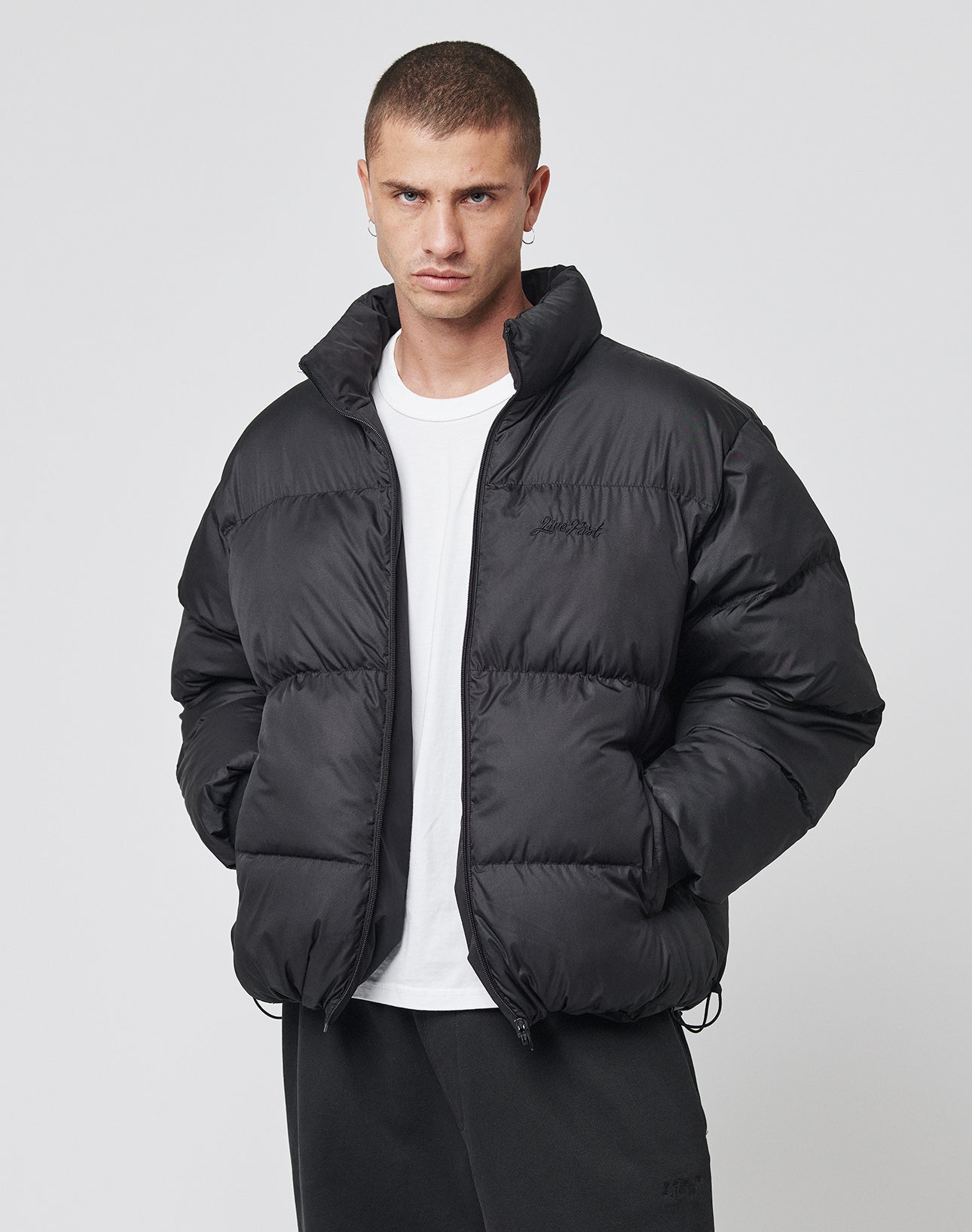A man with short hair wears the LIVE FAST DIE YOUNG LFDY Puffer Jacket, featuring front logo stitching, over a white t-shirt and black pants, standing with hands in his pockets against a plain light background.