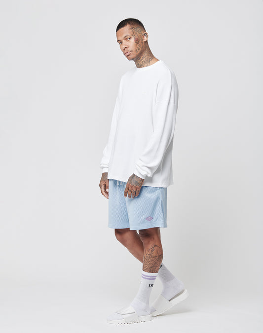 A tattooed person in a relaxed pose stands against a light background, wearing the LIVE FAST DIE YOUNG Basic Waffle Longsleeve in white (Oversize Fit, 100% cotton), paired with light blue shorts, white socks, and sandals.