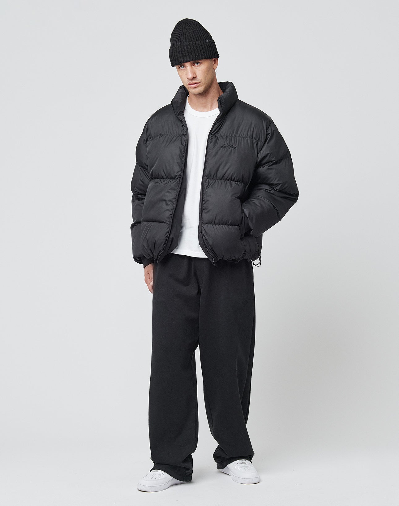 A person in a black LIVE FAST DIE YOUNG LFDY Puffer Jacket with front logo stitching, black beanie, white tee, loose black pants, and white sneakers stands against a plain white background with hands in jacket pockets.