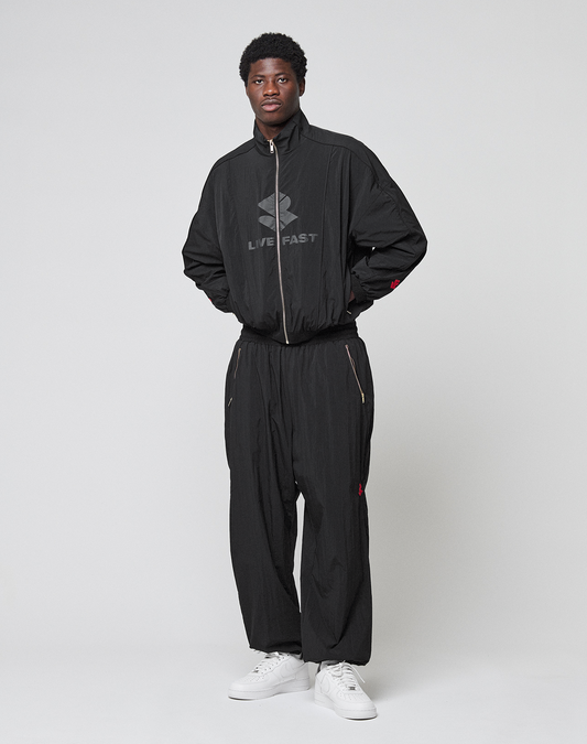 A man poses against a white background wearing LIVE FAST DIE YOUNG’s LFDY Dealership Trackpants and matching black jacket with LIVE FAST branding. The tracksuit has a wide fit, elastic waistband, and he pairs it with white sneakers.